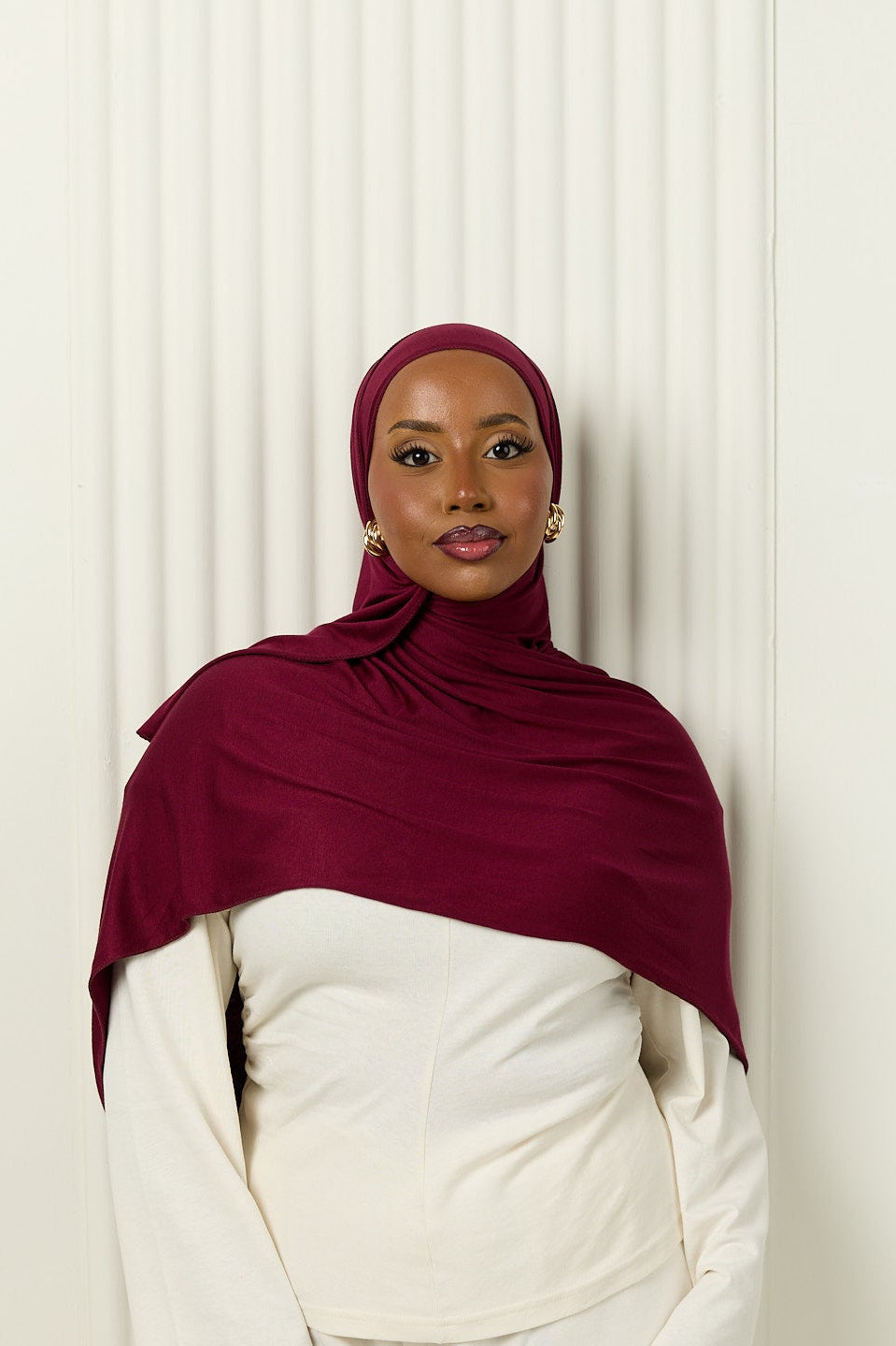 Person wearing a maroon hijab and white outfit against a white curtain background