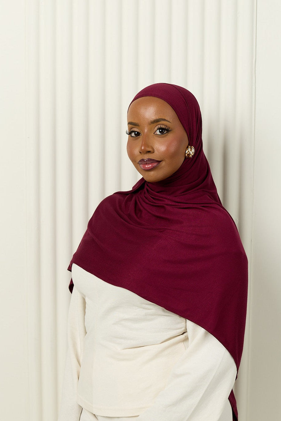 Woman wearing a burgundy hijab and white outfit against a white curtain background